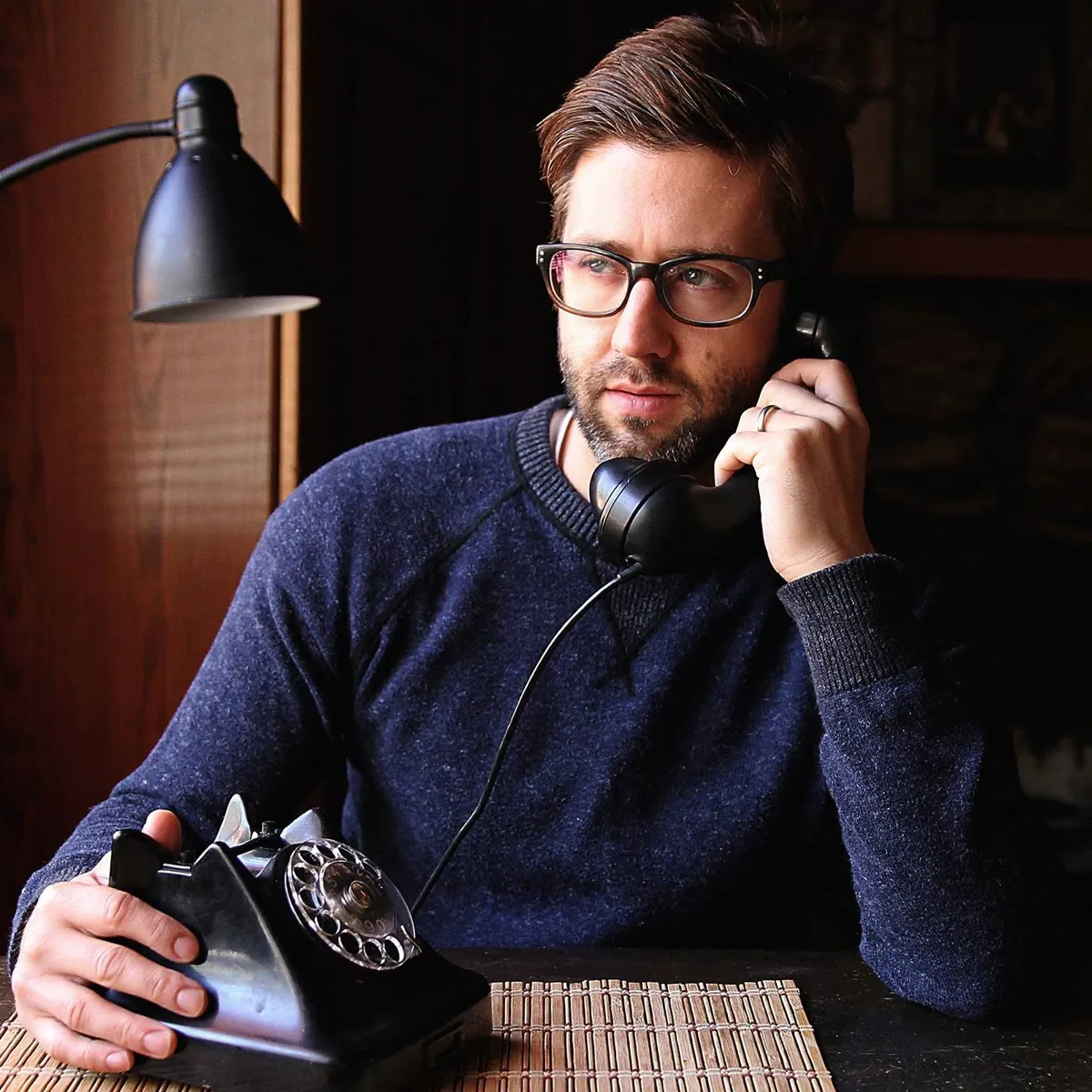 Derek Olson at a desk in a dim room, on the phone, with an old rotary telephone and a desk lamp